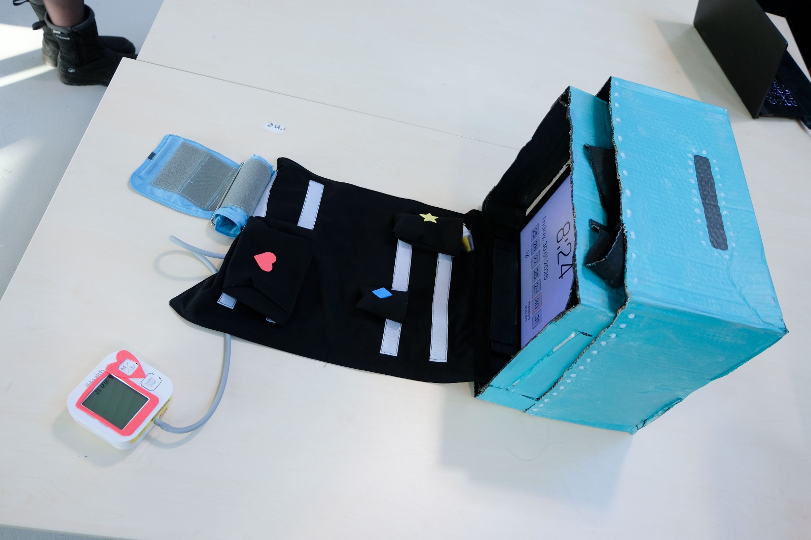 A turquoise cardboard box prototype sits open on a table, revealing a black Velcro-lined interior panel that folds out flat. The panel has reflective strips and several small pockets, each marked with raised foam shapes like hearts, stars, and diamonds to indicate where devices go. A tablet is mounted upright inside the box as a screen, while a digital blood pressure monitor and its cuff extend from one of the foam-marked pockets.  - Image description generated with Be My AI.