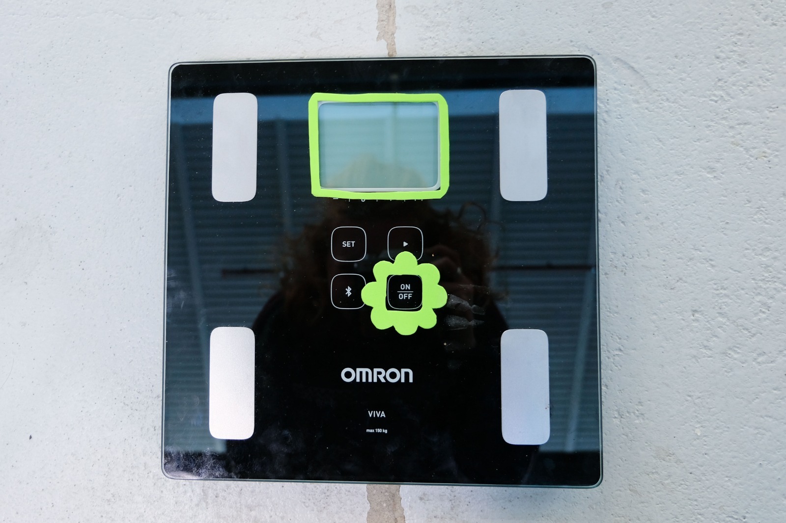 A square Omron Viva digital weighing scale rests on a light-colored floor. The scale has a glossy black glass surface with four vertical metallic footplates near each corner. At the top center is a rectangular display; a thick, bright neon-green foam frame has been stuck around the screen so its edges can be easily traced by touch. Below the display are four touch buttons arranged in a diamond pattern and labeled SET, up arrow, down arrow, and a star-like symbol. Over the central ON/OFF button sits a raised foam cutout shaped like a simple flower, also in neon green, making the power control stand out tactually from the smooth glass surface. The OMRON logo and “VIVA” label are printed in white near the bottom. - Image description generated with Be My AI.