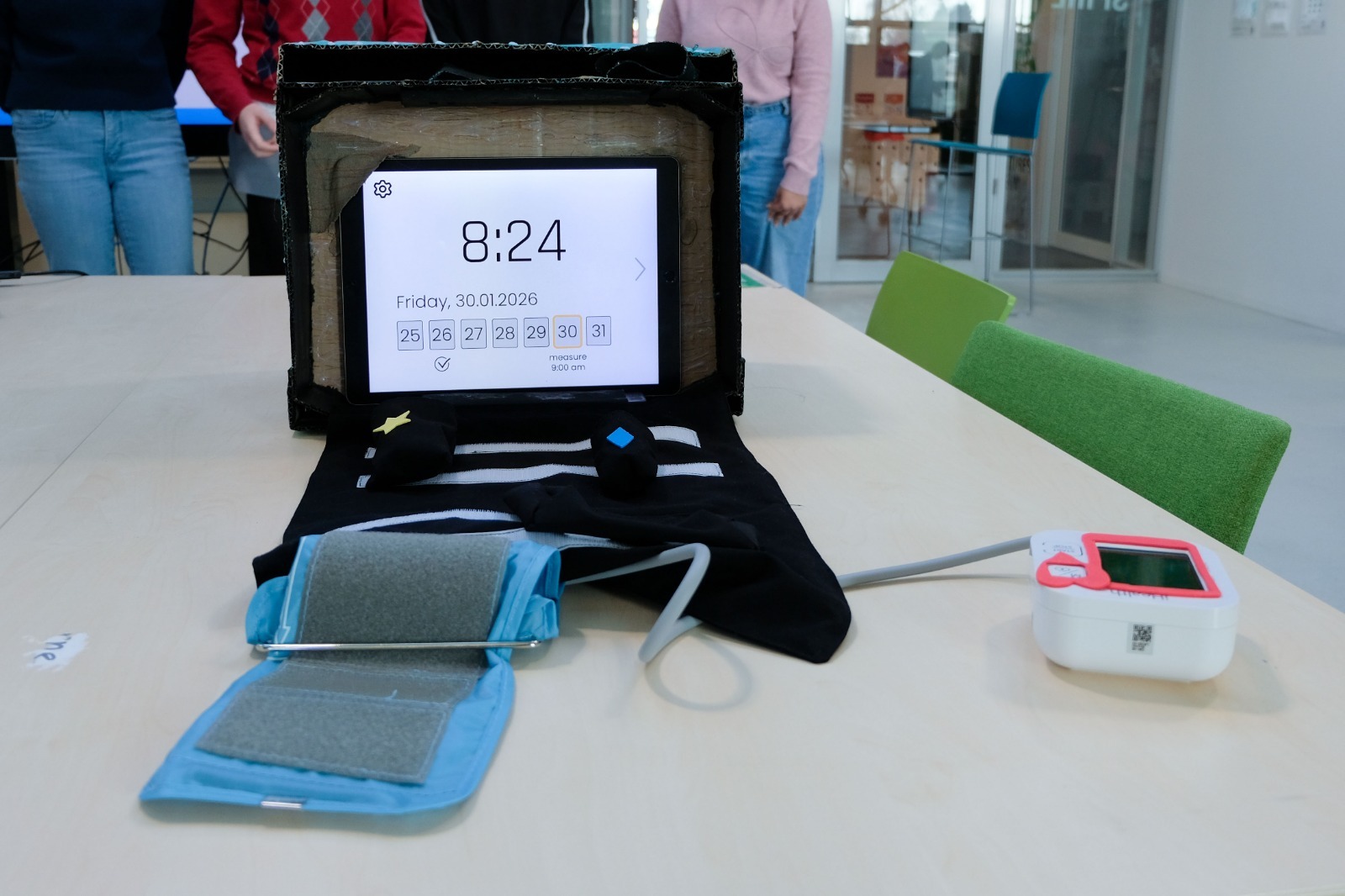 A low-fidelity prototype of a home health toolkit is placed on a long, light wooden table in a bright room. At the center is an open rectangular cardboard box standing on its short edge, with rough, unfinished brown cardboard visible inside and black tape reinforcing the corners. Mounted inside the opening is a tablet in landscape orientation, used as a digital companion screen. The interface shows a minimalist white background with a small gear icon in the top left, the time “8:24” in very large bold digits, and the text “Friday, 30.01.2026” underneath. Below the date is a row of square buttons for calendar days, from 25 to 31, with the current day highlighted and a label “measure 9:00 am” next to it. - image description generated with Be My AI.