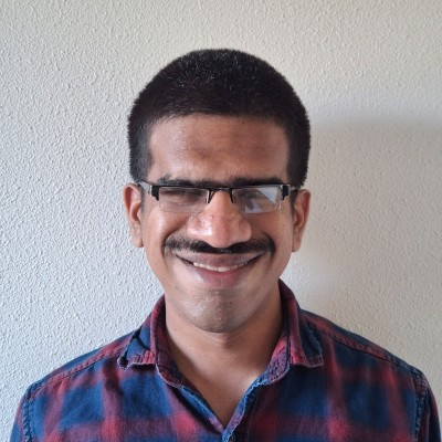 Headshot of Suhas Dharwad. He has short black hair, medium-brown skin, and a neatly trimmed mustache. He is wearing rectangular dark-framed glasses and a casual red-and-blue plaid button-up shirt with the collar open. Suhas is smiling warmly against a plain light-colored background.